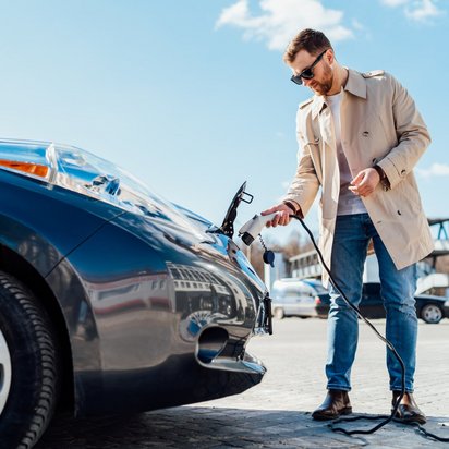 Mann im Trenchcoat steht mit einem Ladekabel in der Hand vor seinem Auto