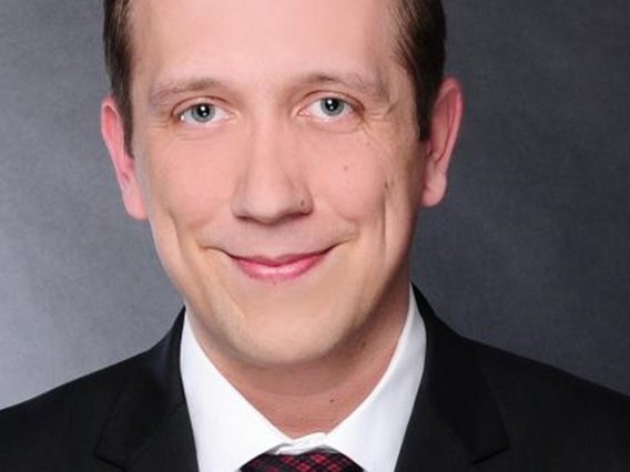 Person mit kurzem hellbraunem Haar trägt einen dunklen Anzug, ein weißes Hemd und eine rot gemusterte Krawatte. Die Person steht vor einem dunkelgrauen, neutralen Hintergrund und lächelt leicht.