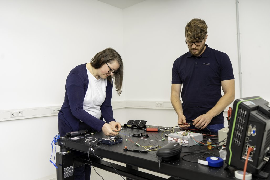 Zwei Personen stehen an einem Tisch, der mit verschiedenen elektronischen Geräten und Kabeln ausgestattet ist. Eine Person trägt ein dunkelblaues T-Shirt mit dem Aufdruck "TÜVIT", die andere Person trägt einen weißen Pullover mit einer blauen Strickjacke. Auf dem Tisch befinden sich Werkzeuge, Kabel und technische Geräte. Im Hintergrund sind weiße Wände und Steckdosen zu sehen.