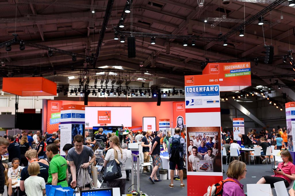 Eine große Messehalle mit orangefarbenen Ständen und Bannern. Im Vordergrund sind Besucher zu sehen, die sich an verschiedenen Ausstellungsständen aufhalten. Ein großes Banner mit der Aufschrift "IDEENFANG" ist sichtbar.