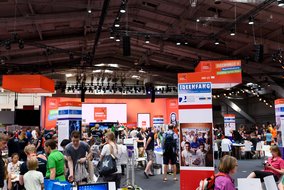 Eine große Messehalle mit orangefarbenen Ständen und Bannern. Im Vordergrund sind Besucher zu sehen, die sich an verschiedenen Ausstellungsständen aufhalten. Ein großes Banner mit der Aufschrift "IDEENFANG" ist sichtbar.