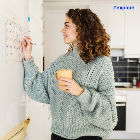Eine Person steht in einer Küche und markiert ein Datum auf einem Wandkalender. Sie trägt einen gemütlichen Strickpullover und hält eine gelbe Tasse in der Hand. Der Hashtag "#explore" ist oben rechts im Bild zu sehen.