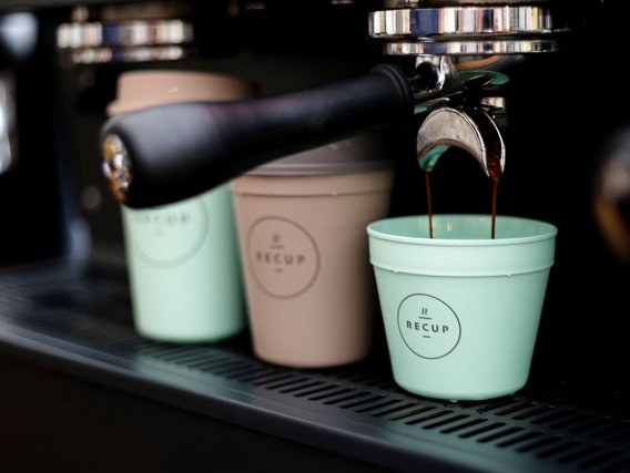 Espressomaschine mit schwarzem Siebträger, aus dem Kaffee in eine hellgrüne RECUP-Tasse fließt, daneben stehen weitere bunte Mehrwegbecher auf dem Metallrost.
