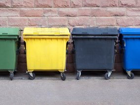 Vier farbige Müllcontainer (grün, gelb, grau und blau) in einer Reihe vor einer rosa Backsteinmauer, für Mülltrennung und Recycling.