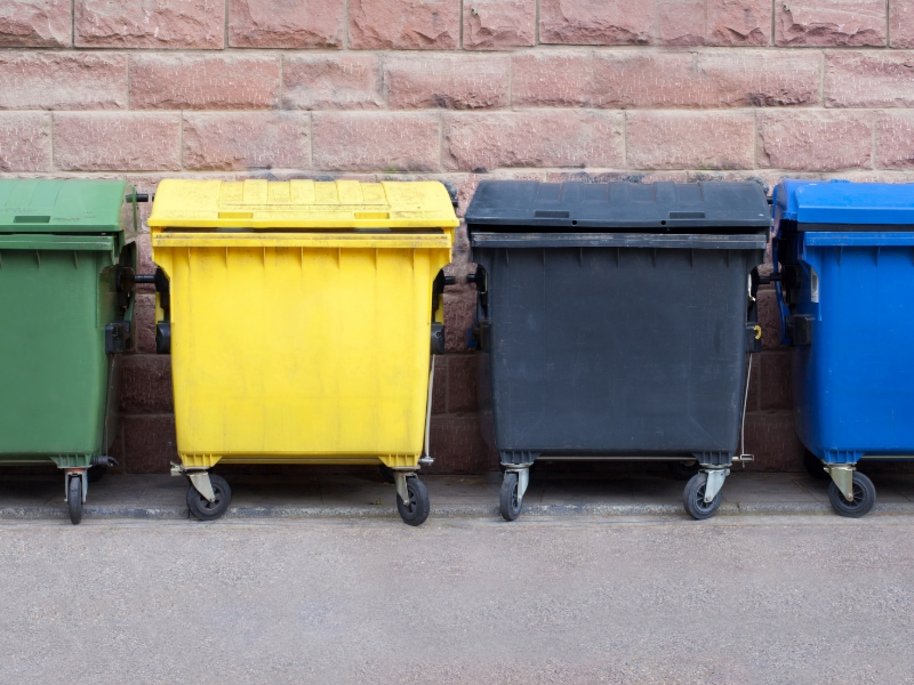 Vier farbige Müllcontainer (grün, gelb, grau und blau) in einer Reihe vor einer rosa Backsteinmauer, für Mülltrennung und Recycling.