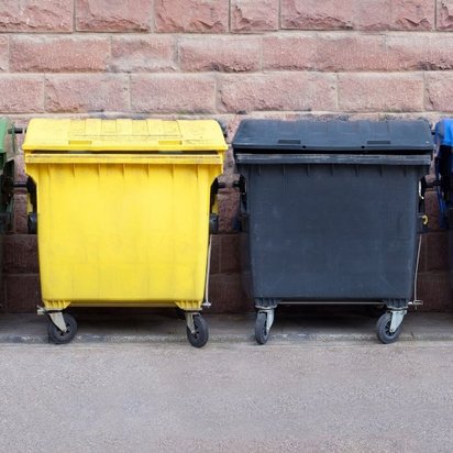 Vier farbige Müllcontainer (grün, gelb, grau und blau) in einer Reihe vor einer rosa Backsteinmauer, für Mülltrennung und Recycling.