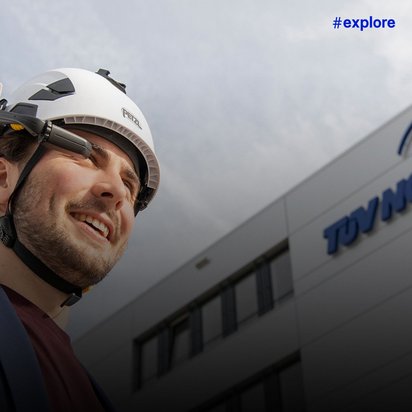 Ein lächelnder Mann mit weißem Schutzhelm und Schutzbrille steht vor einem TÜV NORD Firmengebäude. Der Himmel im Hintergrund ist bewölkt und grau.