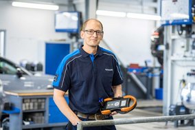 TÜV NORD Mitarbeiter mit Prüfgerät in einer modernen Werkstatt, im Hintergrund Werkstattausrüstung und ein Auto.