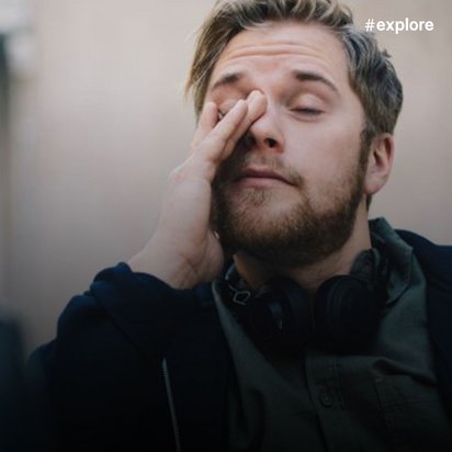 Ein müder junger Mann mit braunen Haaren, Bart und dunkelblauem Sweatshirt, der sich die Augen reibt.