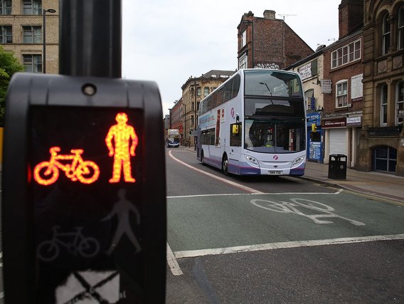 Eine Fußgängerampel zeigt rote Symbole für Fahrrad und Person, während im Hintergrund ein weißer Doppeldeckerbus durch eine britische Stadtstraße fährt. Die Ampel steht im Vordergrund scharf, der Bus ist leicht unscharf im Hintergrund zu sehen.