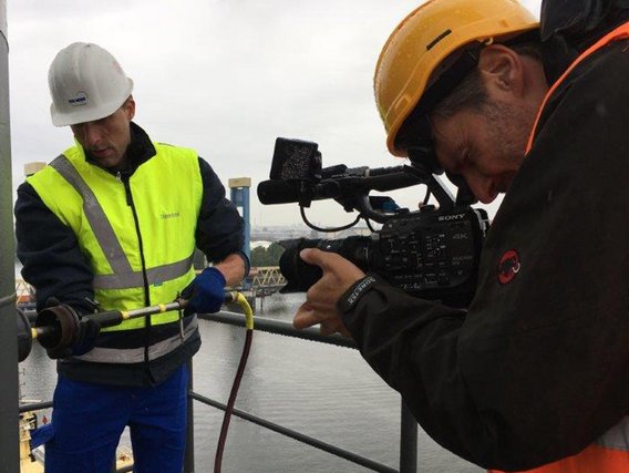 Zwei Arbeiter mit Schutzhelmen auf einer Baustelle, einer in gelber Sicherheitsweste arbeitet an einem Rohr, während der andere mit einer professionellen Videokamera filmt.