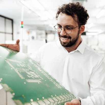 Eine Person hält eine große Leiterplatte in den Händen und überprüft deren Bauteile in einer technischen Werkstatt.