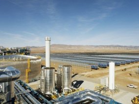 Eine große Solaranlage in einer trockenen Wüstenregion mit industriellen Anlagen im Vordergrund. Silberfarbene Tanks und Rohre sind sichtbar, während im Hintergrund Reihen von Solarkollektoren unter blauem Himmel zu sehen sind.