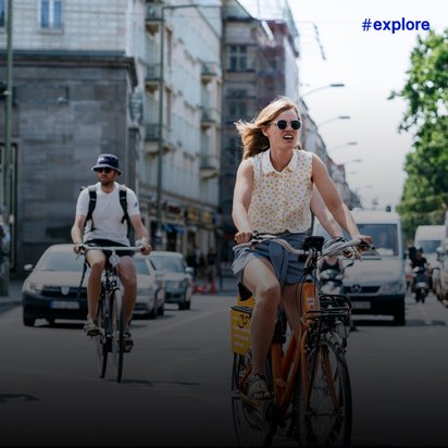 Drei Radfahrer in sommerlicher Kleidung auf einer städtischen Straße; eine lächelnde Frau mit Sonnenbrille und gelbem Top vorne, dahinter ein Mann mit Hut und eine Frau in grauem Shirt, parallel zum Autoverkehr.