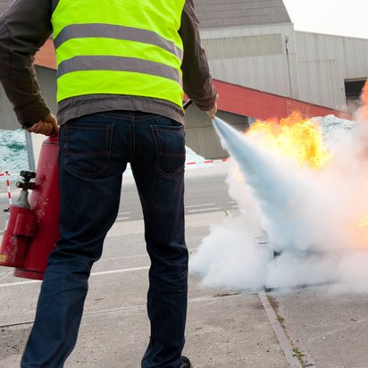 Löschübung im Freien zur Schulung im Brandschutzverhalten. Person mit Warnweste löscht bei einer Übung im Freien ein offenes Feuer mit einem Feuerlöscher.