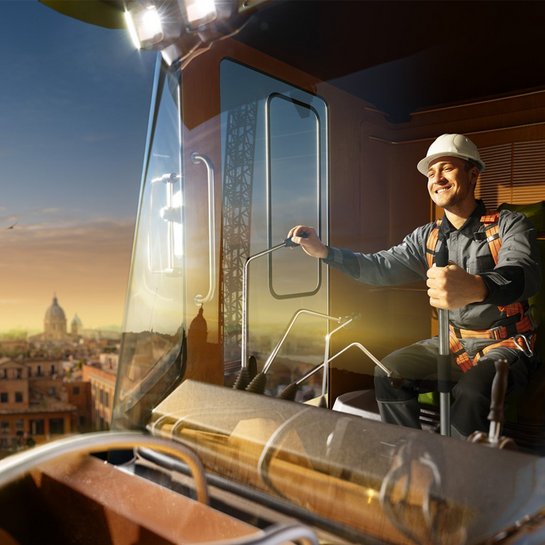  Ein Kranführer sitzt in der Kabine eines Turmkrans und steuert die Maschine, während im Hintergrund eine Stadtlandschaft bei Sonnenuntergang zu sehen ist. Die Szene vermittelt ein Gefühl von Höhe und Kontrolle über die urbanen Bauarbeiten.