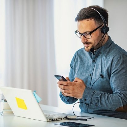 Ein Mann mit Brille und Headset in einem blauen Hemd schaut auf sein Smartphone, während er an einem Tisch mit Laptop und Wasserglas sitzt.