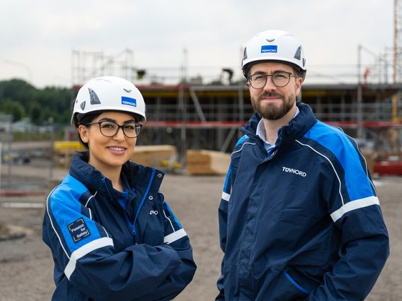 Zwei BauingenieurInnen mit Schutzhelmen und blauer Arbeitskleidung stehen vor einer Baustelle und lächeln in die Kamera.