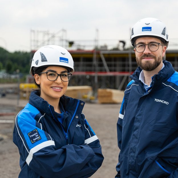 Zwei BauingenieurInnen mit Schutzhelmen und blauer Arbeitskleidung stehen vor einer Baustelle und lächeln in die Kamera.