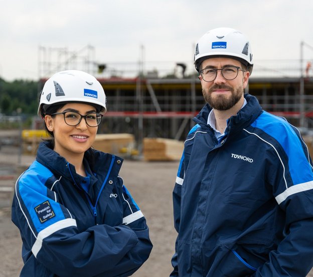 Zwei BauingenieurInnen mit Schutzhelmen und blauer Arbeitskleidung stehen vor einer Baustelle und lächeln in die Kamera.