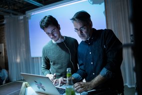 Zwei Personen, die gemeinsam an einem Laptop arbeiten, in einem abgedunkelten Raum mit blauer Beleuchtung.