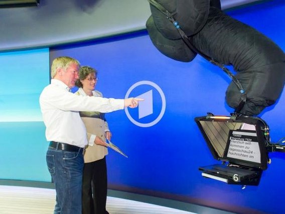 Ein Nachrichtenstudio mit zwei Personen, die vor einem blauen Hintergrund mit Weltkarte stehen. Sie zeigen auf einen Teleprompter, während eine große Kamera mit Textanzeige auf sie gerichtet ist.