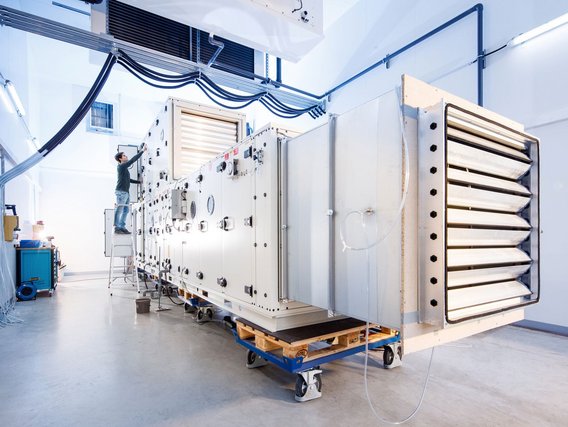 Ein Techniker auf einer Leiter arbeitet an einer großen weißen Lüftungsanlage mit mehreren Klappen und Steuerungselementen.
