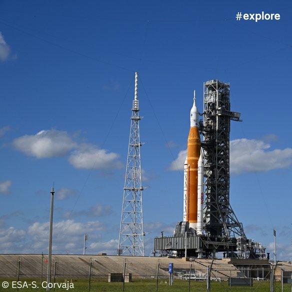 Eine große Rakete steht auf einer Startrampe, umgeben von hohen Türmen und technischen Anlagen. Der Himmel ist klar und blau mit einigen Wolken. Die Szene vermittelt den Eindruck von technologischem Fortschritt und Raumfahrt.