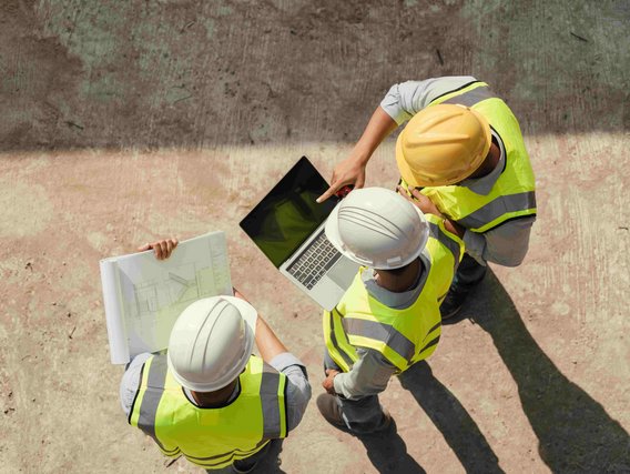 Drei Bauingenieure in Sicherheitswesten und Helmen stehen auf einer Baustelle und betrachten gemeinsam einen Laptop und Baupläne.