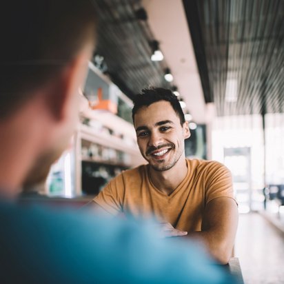 Zwei Personen sitzen in einem stilvollen Café und unterhalten sich. Die Umgebung ist modern gestaltet mit einer hohen Decke und einer einladenden Atmosphäre. Eine Person trägt ein orangefarbenes T-Shirt und sitzt gegenüber der anderen Person. Im Hintergrund sind Regale und eine Glastür zu sehen, die Tageslicht hereinlässt.