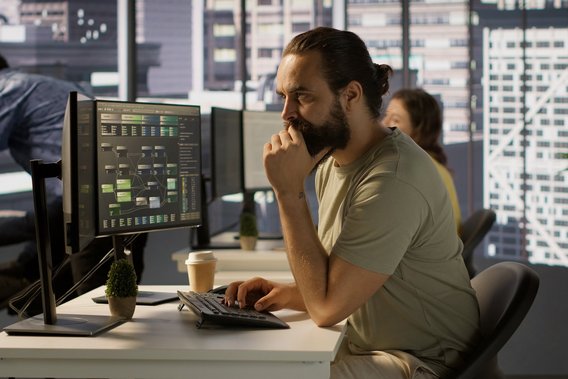 Mann mit Bart arbeitet fokussiert am Computer mit Datenvisualisierung auf dem Monitor im modernen Büro