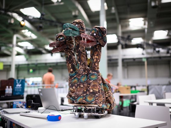 Ein dekorativer Roboter mit mehreren "Schlangenköpfen", gestaltet mit schlangenartigem Muster in braun-grün. Er steht auf einer weißen Tischfläche in einer großen Halle und hat drei Köpfe mit augenartig gestalteten Elementen.