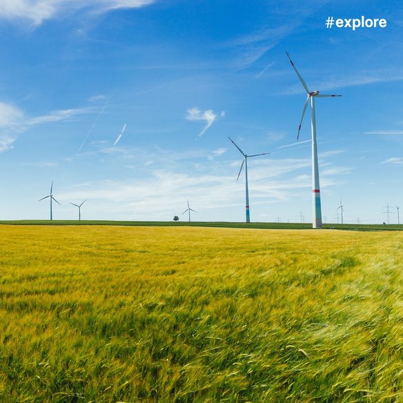 Eine Reihe von Windkraftanlagen steht in einem weiten Feld mit grünem Gras unter einem klaren blauen Himmel. Der Hashtag "#explore" ist oben rechts im Bild zu sehen.
