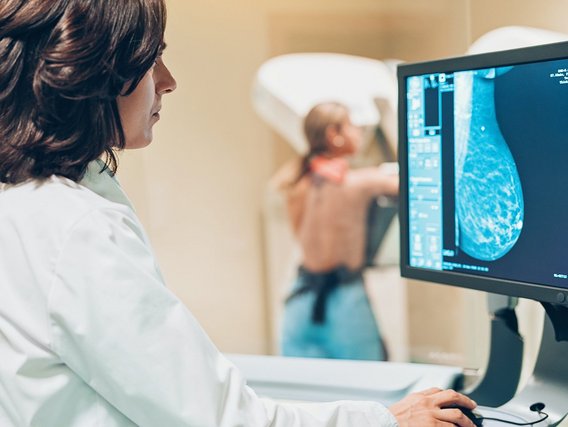 Eine Ärztin in weißem Kittel betrachtet eine Mammografie-Aufnahme auf einem Monitor, während eine Patientin im Hintergrund unscharf zu sehen ist. Die Szene zeigt eine typische radiologische Untersuchungssituation in der Brustkrebsvorsorge.