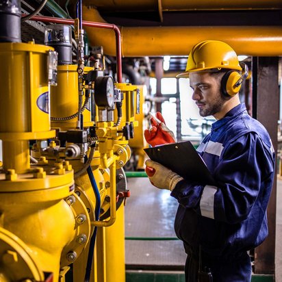 Techniker mit Helm und Gehörschutz überprüft gelbe Industrieanlage mit einem Klemmbrett in der Hand.
