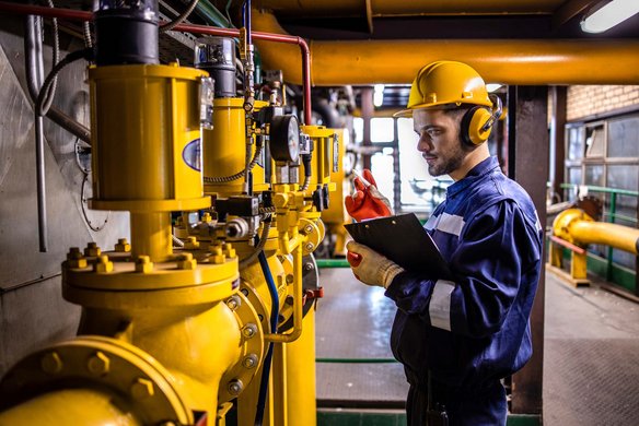  Techniker mit Helm und Gehörschutz überprüft gelbe Industrieanlage mit einem Klemmbrett in der Hand.
