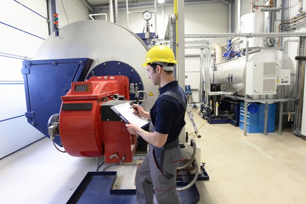 Techniker mit Schutzhelm prüft Industrieanlage mit roter Maschine und macht Notizen auf einem Klemmbrett