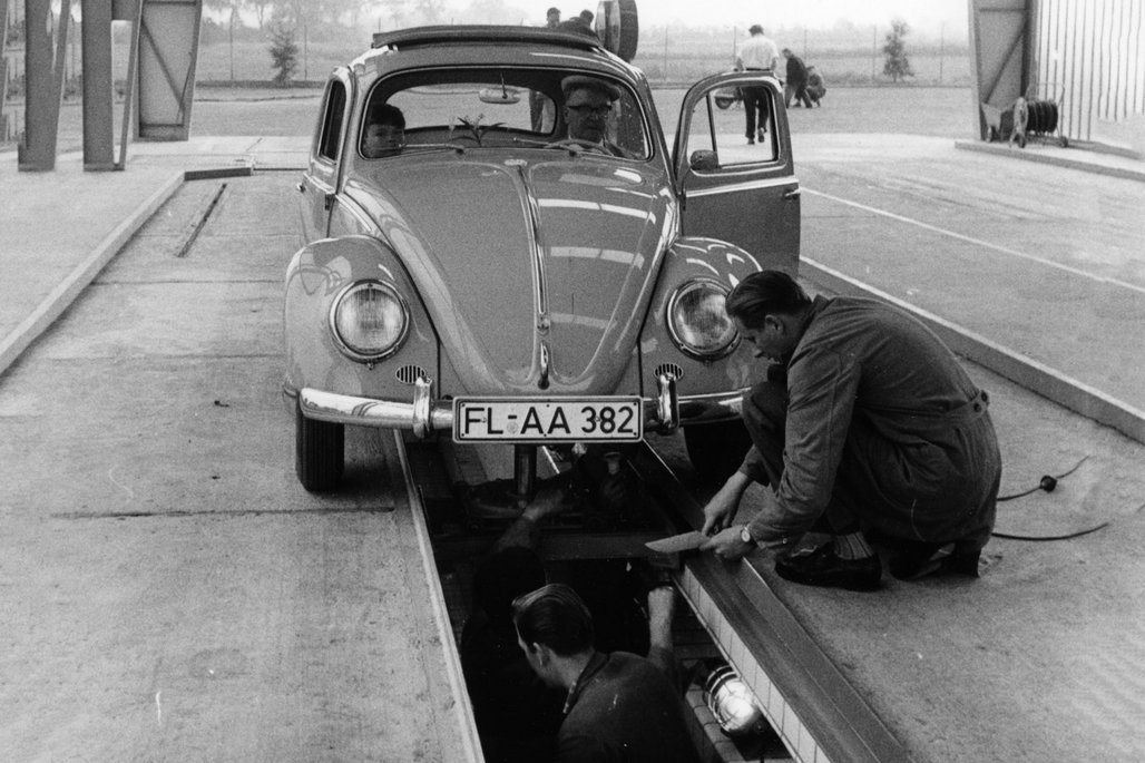Schwarz-weiß Aufnahme eines VW Käfers bei einer Abgasuntersuchung auf einer Hebebühne in den 1960er Jahren.