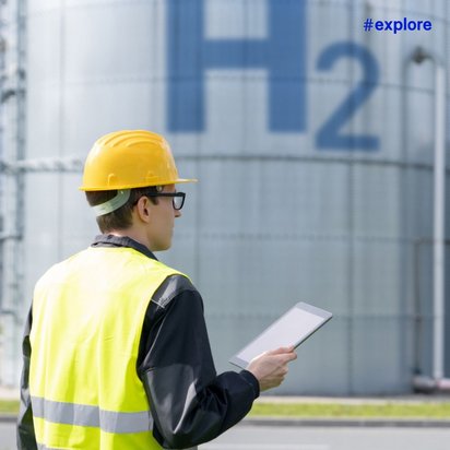 Ein Arbeiter mit gelbem Schutzhelm und Warnweste steht vor großen Wasserstofftanks. Auf einem Tank ist deutlich "H2" zu sehen, während der Arbeiter ein Tablet in der Hand hält.