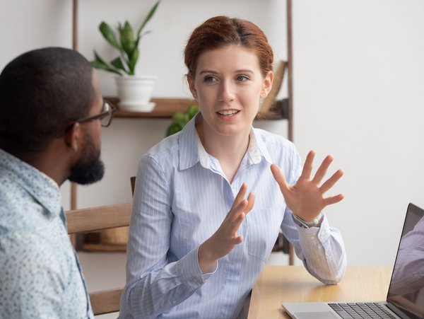 Zwei Personen in einem Gespräch am Tisch, eine Person erklärt etwas mit Gesten, während sie auf ein Laptop schaut