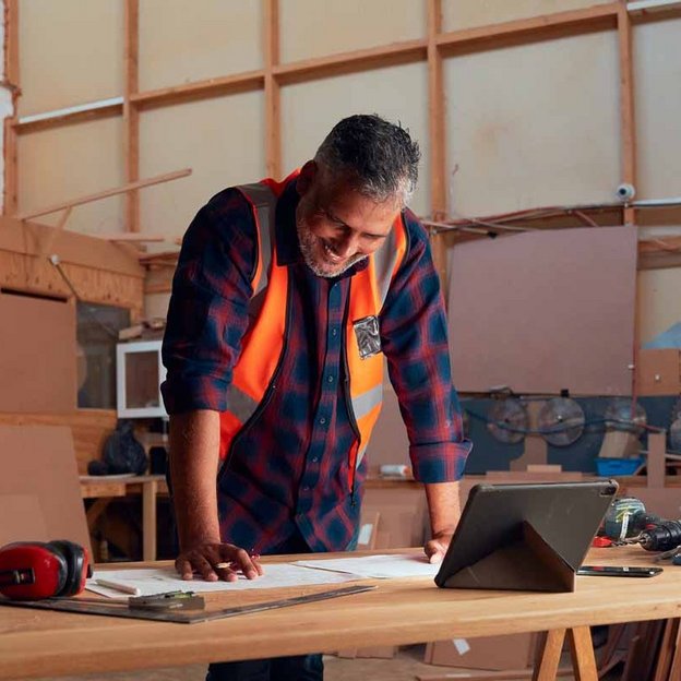  Ein Mann in einer orangefarbenen Sicherheitsweste steht an einem Tisch in einer Werkstatt und betrachtet Pläne auf einem Tablet. Die Szene zeigt Planung und Weiterbildung im handwerklichen Bereich.