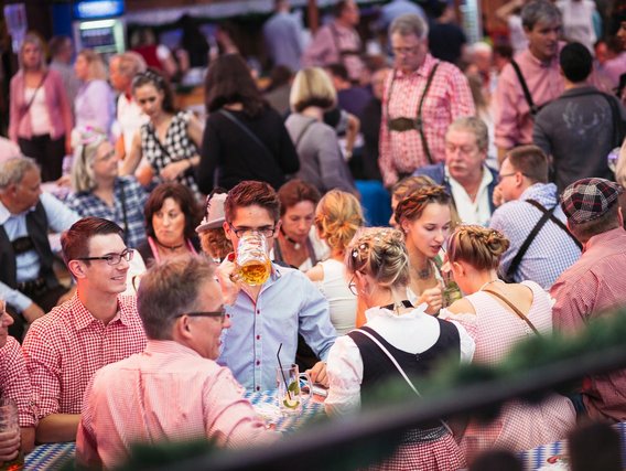 Blick auf eine überfüllte Festveranstaltung mit Personen in traditioneller Kleidung, viele in karierter Tracht, die an Biertischen sitzen und gesellig feiern.
