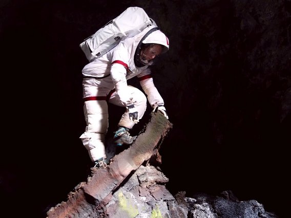 Astronaut in weißem Anzug sammelt Gesteinsproben in einer dunklen Höhle mit gelblich-grünen Mineralablagerungen.