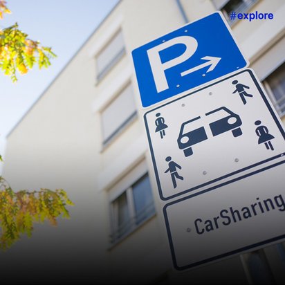 Ein blaues Parkplatzschild mit weißem "P" und Pfeil, darunter ein weißes Zusatzschild mit Carsharing-Symbol und der Aufschrift "CarSharing". Das Schild steht vor einem Gebäude mit herbstlich gefärbten Bäumen.