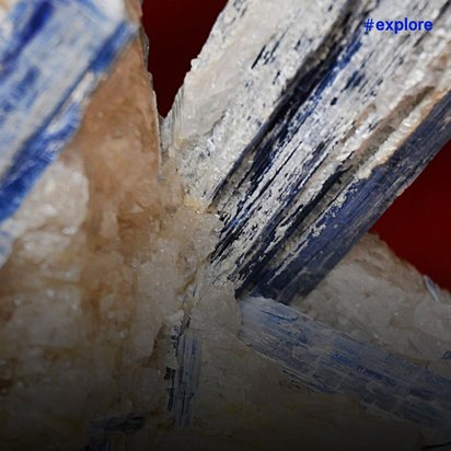 Langgestreckte, prismatische Kristalle in Weiß und Blau mit streifenartigen Mustern, teilweise durchscheinend und von einem rötlichen Hintergrund umgeben.