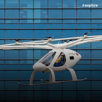 Ein weißes elektrisches Flugtaxi mit Kennzeichen "D-HIDVO" schwebt vor einer blauen Glasfassade eines modernen Gebäudes.