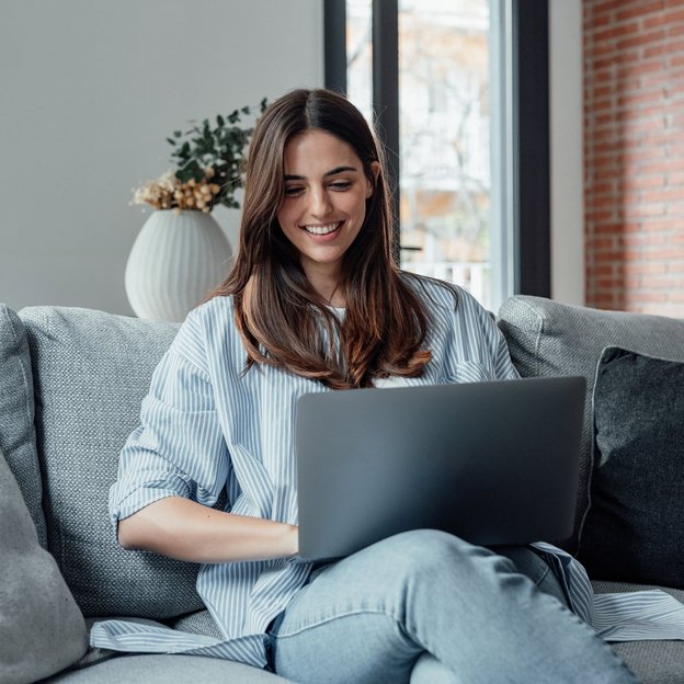 Junge Frau sitzt auf einem Sofa und arbeitet lächelnd an einem Laptop