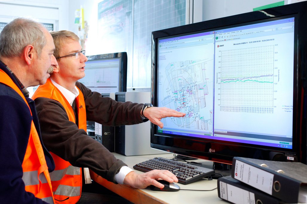 In einem Büro sitzen zwei Personen vor einem großen Computerbildschirm. Eine Person zeigt auf den Bildschirm, der technische Zeichnungen und Diagramme anzeigt. Beide tragen orangefarbene Warnwesten. Auf dem Schreibtisch liegen mehrere Aktenordner, und im Hintergrund sind weitere Computer und technische Ausrüstungen zu sehen.