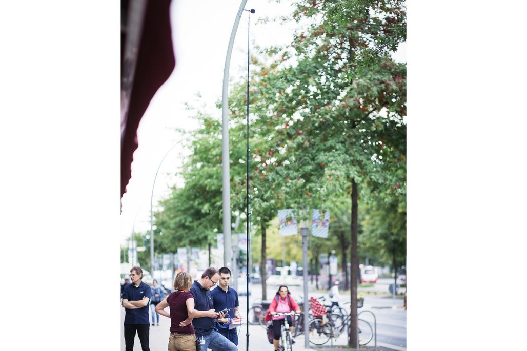 Eine kleine Gruppe von Personen steht auf einer Fußgängerzone vor Geschäften, während eine Frau mit Headset spricht und technisches Equipment für Aufnahmen aufgebaut ist.