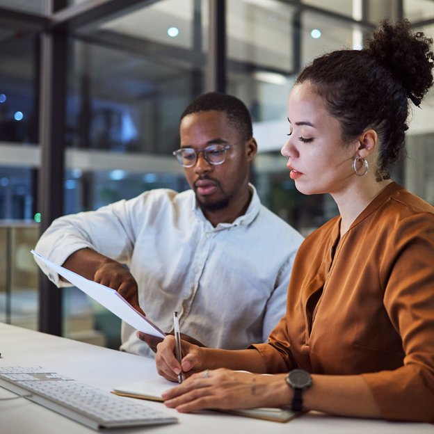 Zwei Kollegen arbeiten abends gemeinsam am Computer und besprechen Unterlagen im modernen Büro